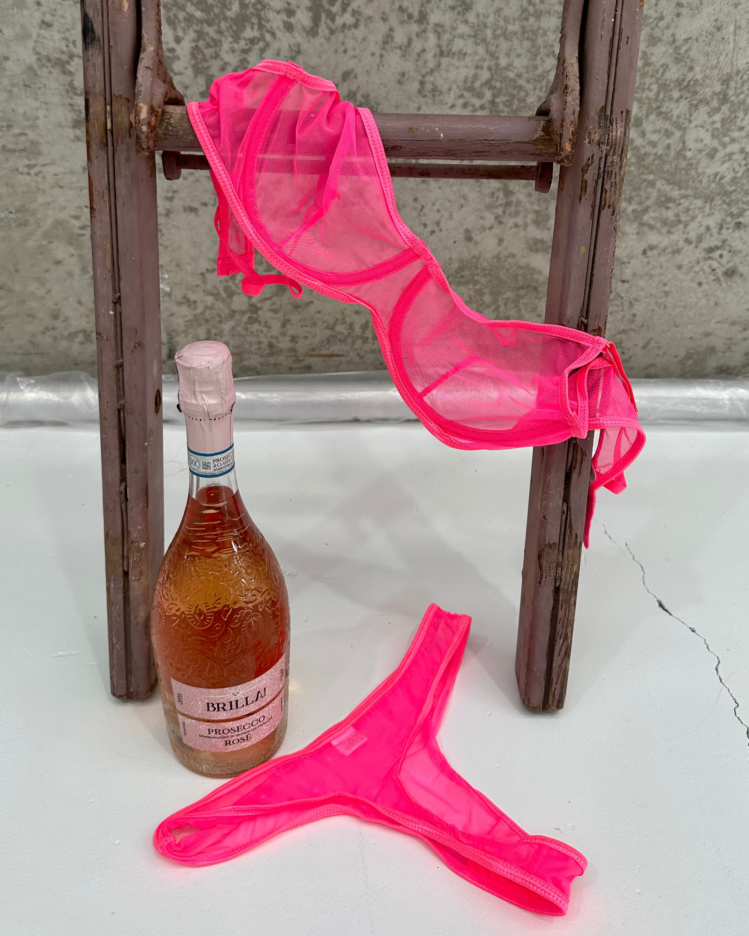 Pink sheer lingerie draped over a wooden chair with a bottle of alcohol on a white surface.